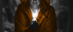 black and white photo of a man in dark cave lit by candle he is holding. His jacket is coloured orange