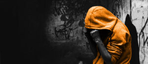 Black and white photo of a man leaning against a graffiti wall with his hands on his face. His hood is up and his hoody is coloured orange
