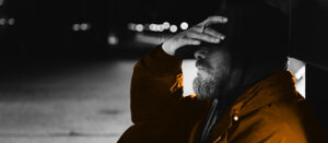 Black and white photo of a man sitting on the ground with his hand over his face. His jacket is coloured orange